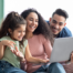 Smiling parents and young daughter sitting together on a couch, looking at a laptop screen.